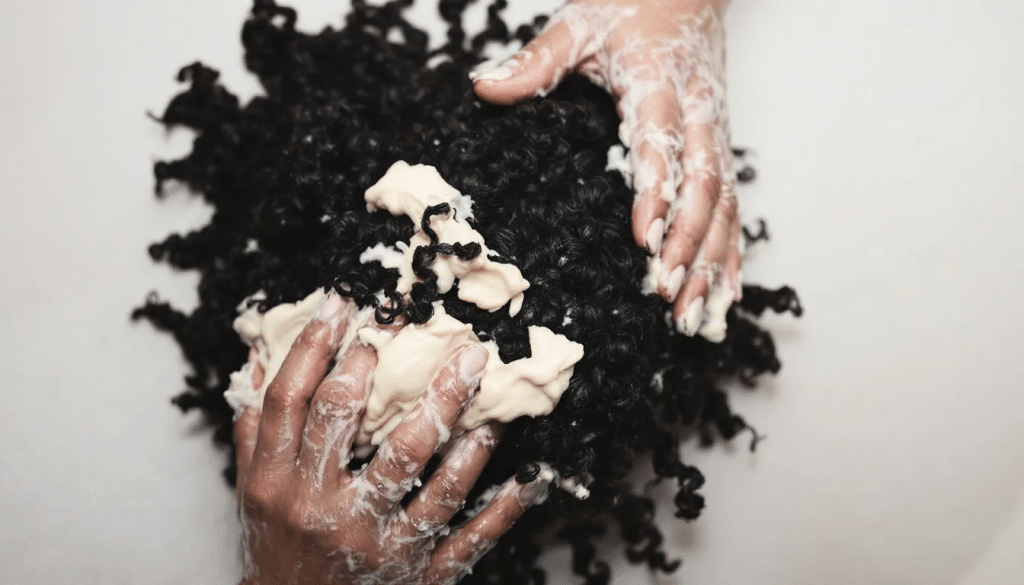 The image shows a stylist's hands applying various styling products to wet curly hair, emphasizing the care and expertise needed to manage natural curls. This scene captures the essence of a curly hair salon, where clients can feel comfortable and embrace their unique hair types.